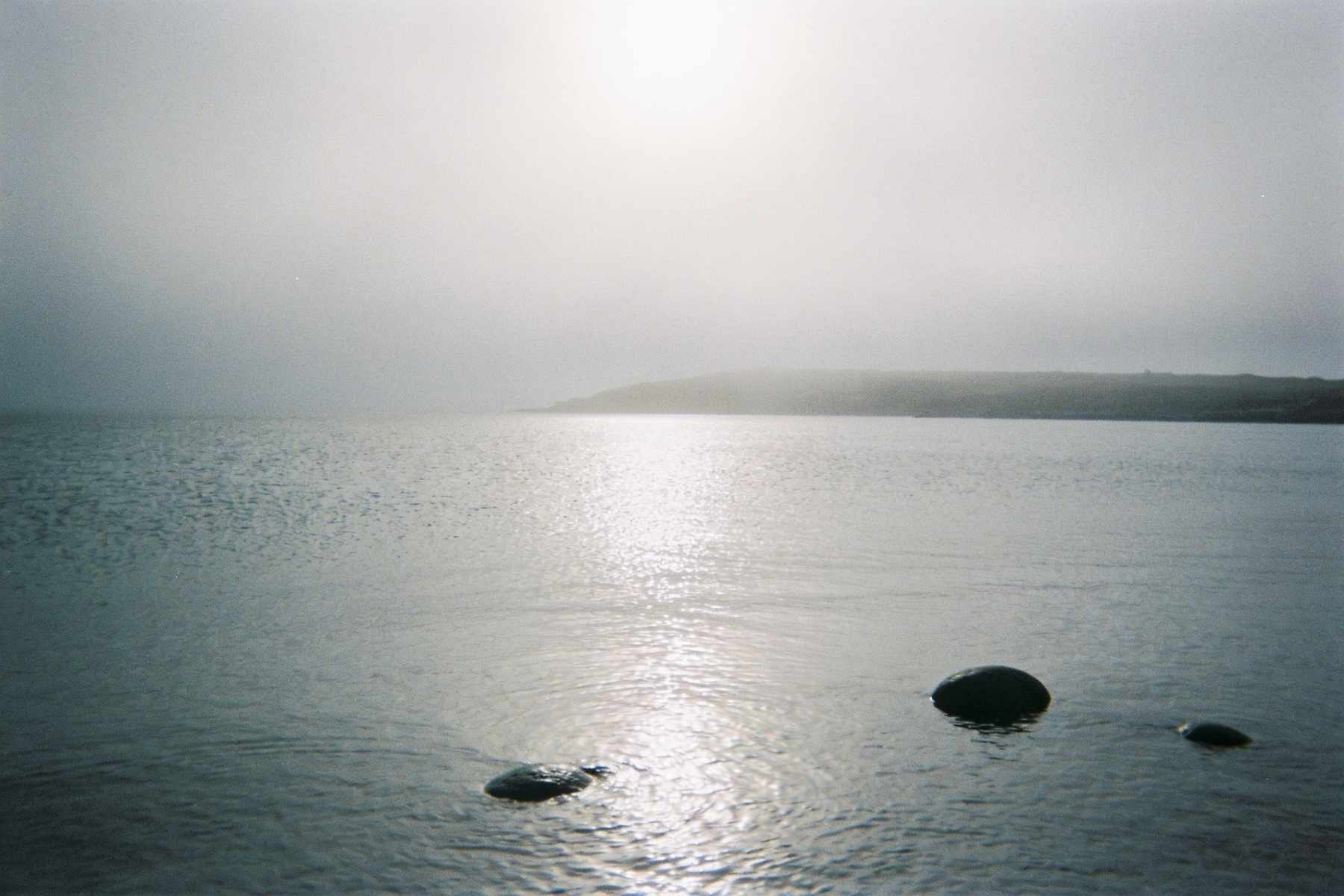 A still, silver sea on a misty winter morning, a few round stones breaking the surface and a faint headland in the fog