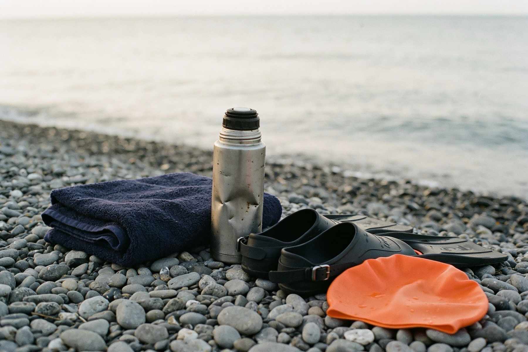 A folded towel, a dented steel flask, neoprene swim socks and a bright orange swim cap on wet grey shingle by a calm sea
