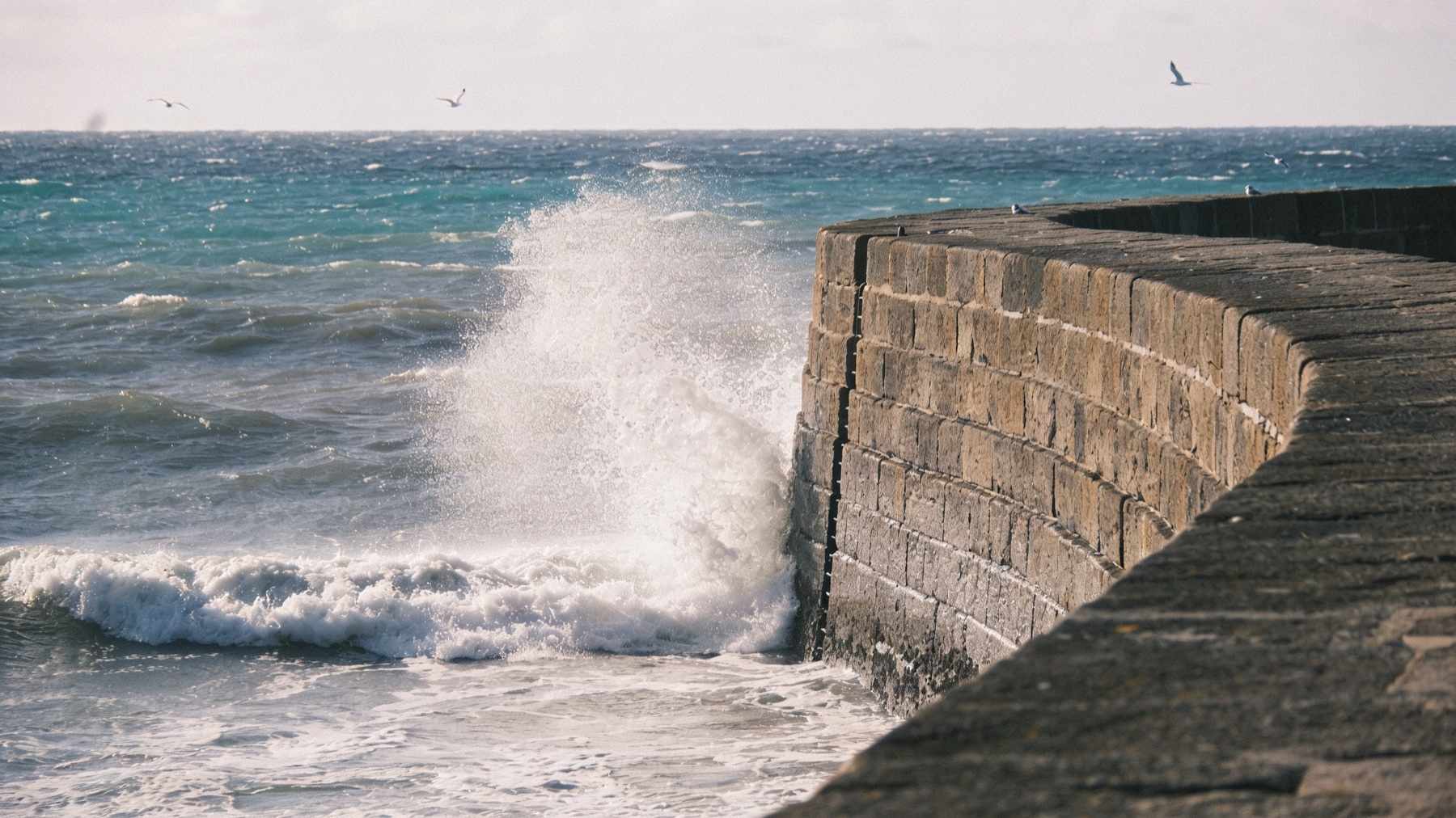 Waves breaking against a curved granite harbour wall on a bright windy day, spray thrown into the air