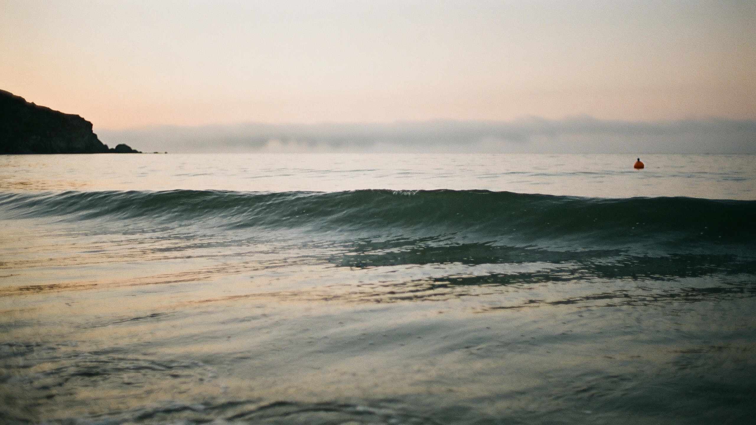 A calm grey-green sea at dawn seen from just above the water, a low swell rolling in, mist on the horizon and a dark headland on the left