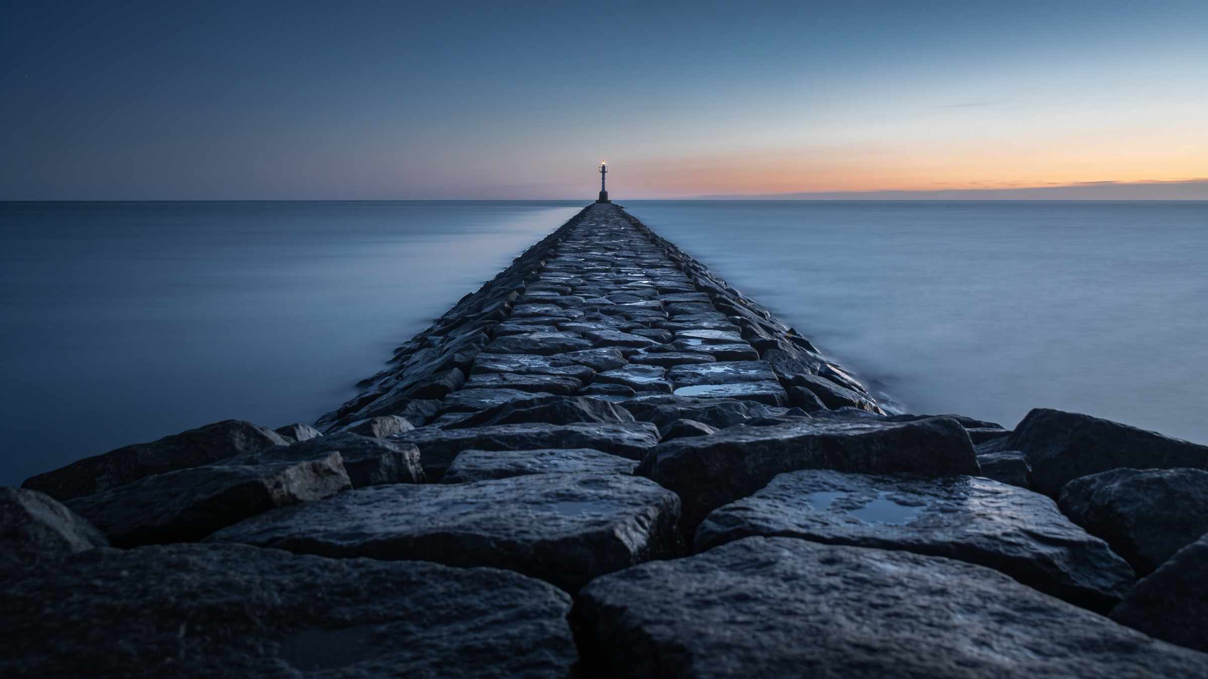 A long stone breakwater running into a flat calm sea at blue hour, a small light at its end and a thin band of orange on the horizon