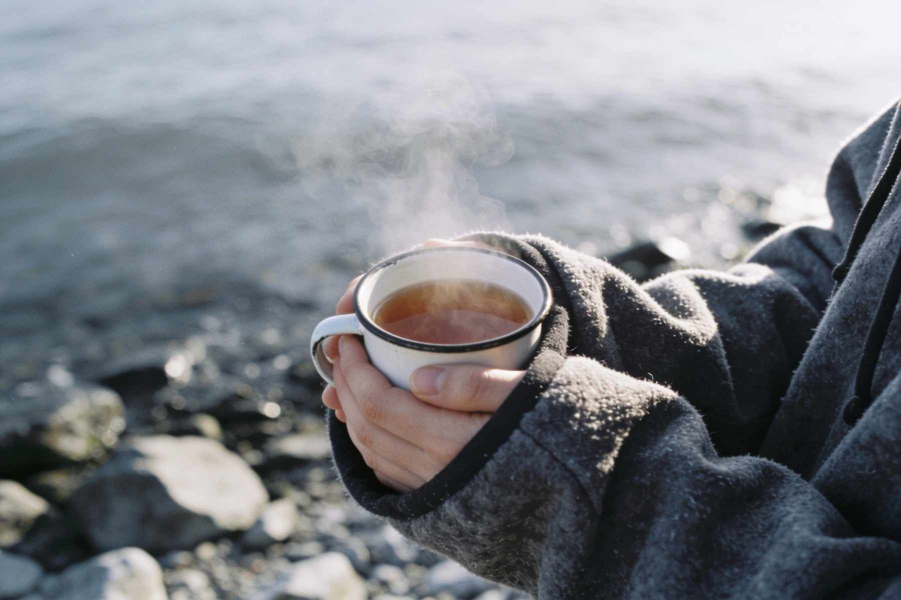 Two cold hands wrapped in the sleeves of a fleece changing robe holding a steaming enamel cup, a rocky shore and grey sea behind